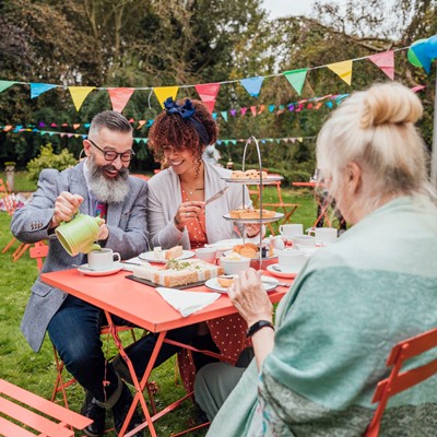 a young mixed race female sitting with a caucasian male and an senior female chatting in a garden, pouring tea, at a garden tea party